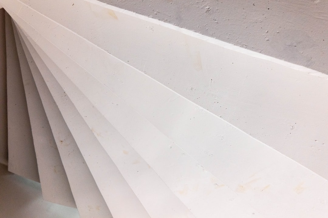 Close-up view of overlapping white panels or sheets arranged in a fanned, diagonal pattern against a light-colored wall, creating an abstract geometric design.