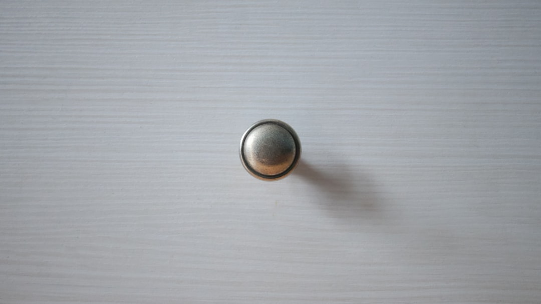 A round, metallic drawer knob is centered on a light-colored, textured wooden surface, casting a faint shadow to the right.