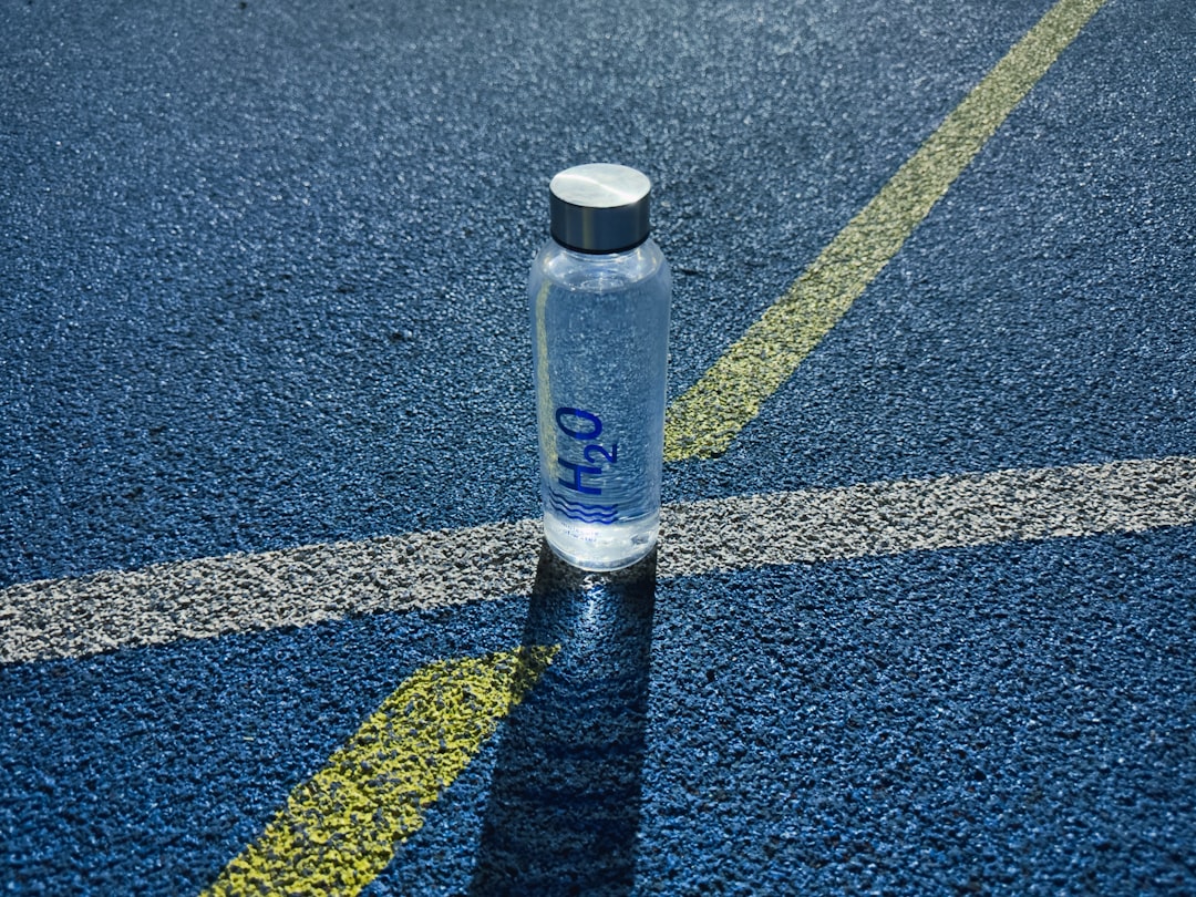 A clear water bottle labeled "H2O" sits on a blue textured track surface, intersected by white and yellow painted lines in sunlight.