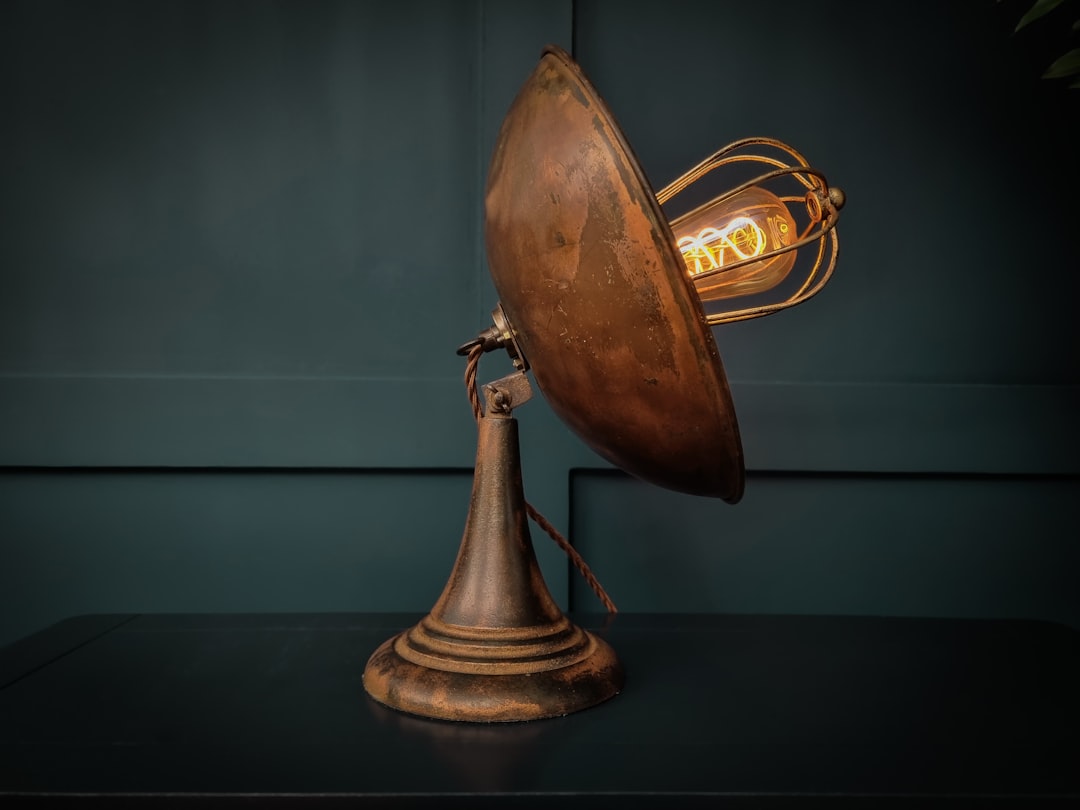A vintage, industrial-style desk lamp with a round, rust-colored metal shade and exposed filament bulb sits on a dark surface against a dark green background.