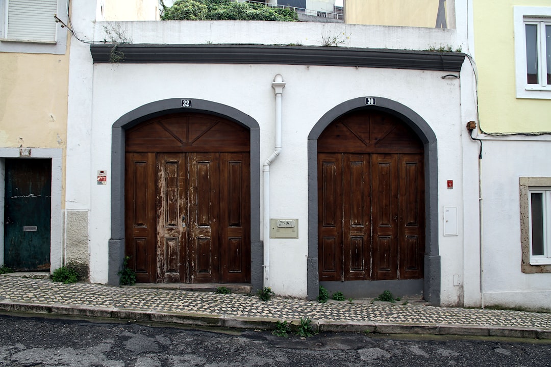 Two large wooden garage doors with arched frames on a white building. The left door shows signs of weathering, while the right is in better condition. Cobblestone pavement and a sloped street are in front.