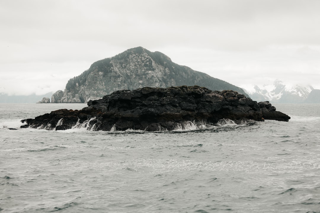 A small, rocky outcrop rises from the ocean with waves splashing against it; a larger forested mountain and cloudy skies are visible in the background.