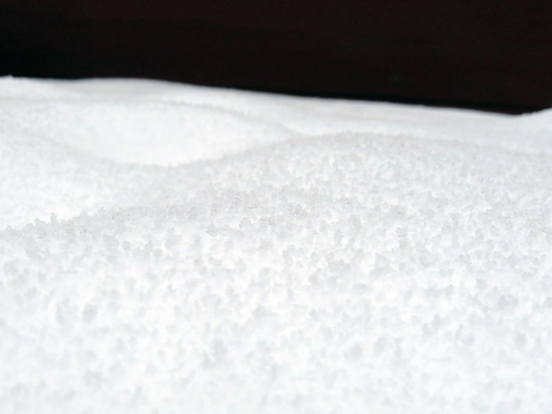 Close-up of a white, uneven surface covered with coarse salt crystals, creating a textured appearance. The background is dark and out of focus.