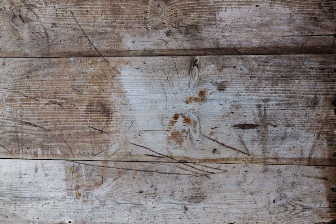 Weathered wooden boards with visible grain, scratches, and stains. The surface appears aged and worn, with various marks and discolorations, giving it a rustic and textured appearance.