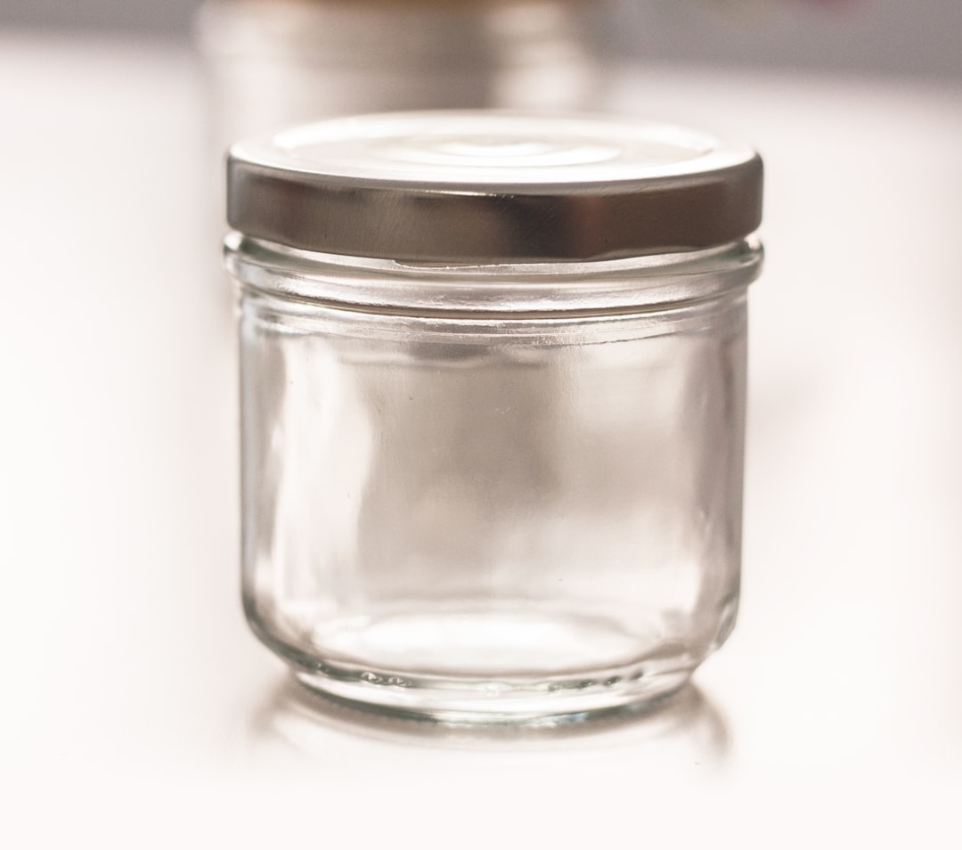 A clear, empty glass jar with a metal lid sits on a white surface. The background is softly blurred, showing another jar out of focus.