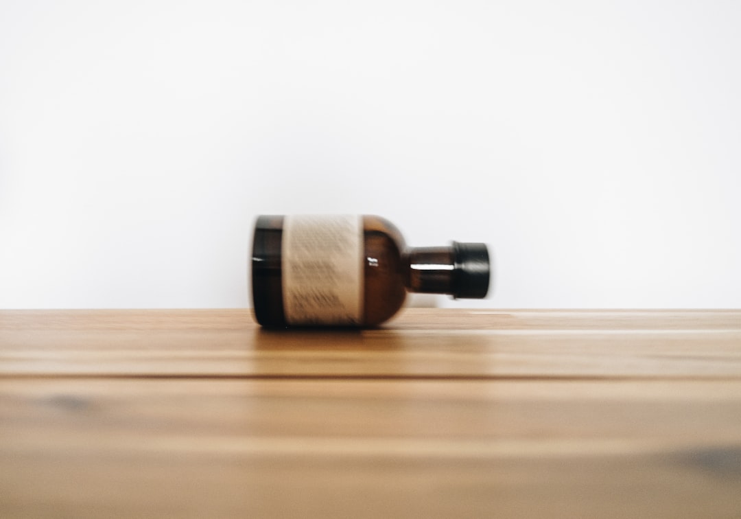 A small brown glass bottle with a black cap lies on its side on a wooden surface against a plain white background. The bottle's label is blurred and unreadable.