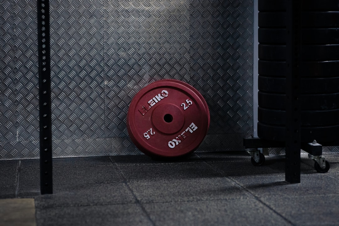 A red Eleiko 25 kg weight plate leans against a metal-textured wall in a gym, with black weight plates stacked on a cart nearby on a rubber gym floor.