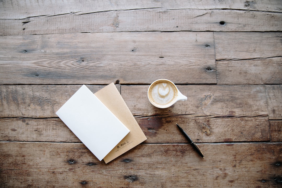 A cup of latte with heart-shaped foam art sits on a rustic wooden table next to an envelope, a white notebook, and a black pen.