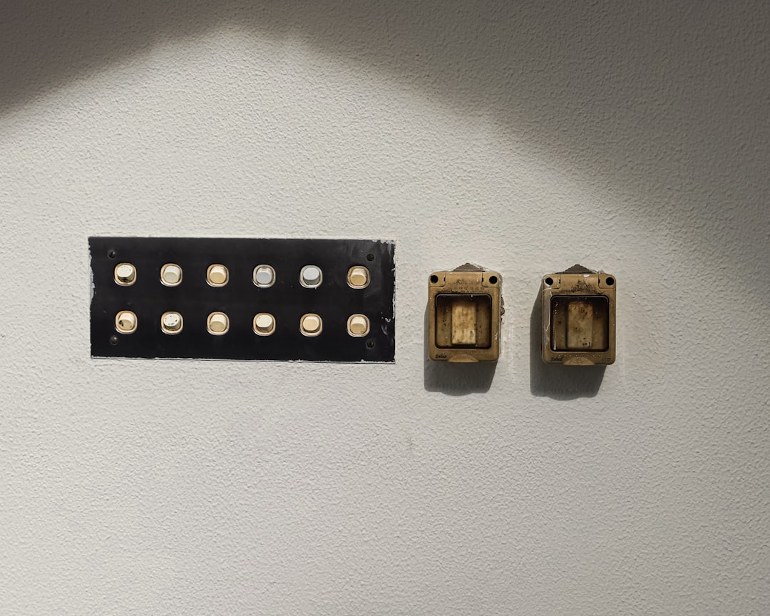 A white wall with a metal panel featuring twelve round white buttons arranged in two rows, next to two vintage, square, brass-colored electrical switches. Light casts a shadow across the top of the image.