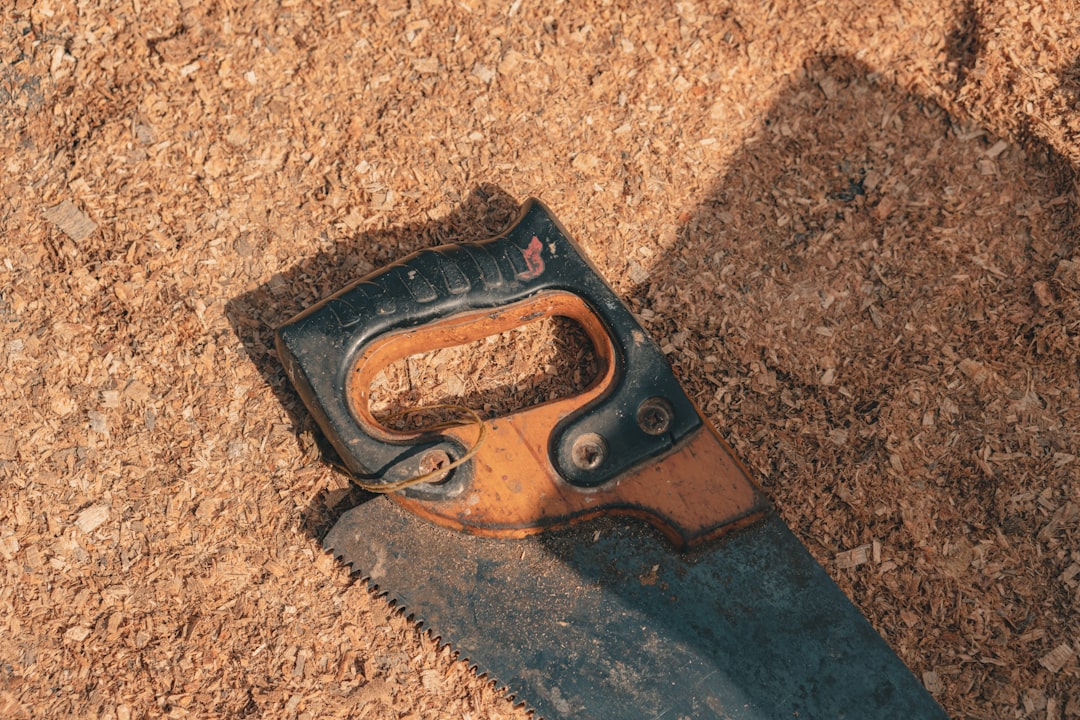 A close-up of a hand saw with a black and orange handle lying on a surface covered in sawdust, casting a shadow to the right.