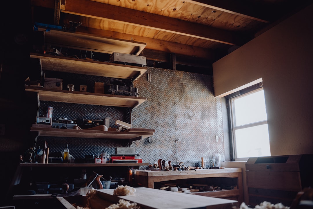 A woodworking shop with wooden shelves holding tools and supplies, a bench with woodworking tools, and sunlight streaming in through a window.