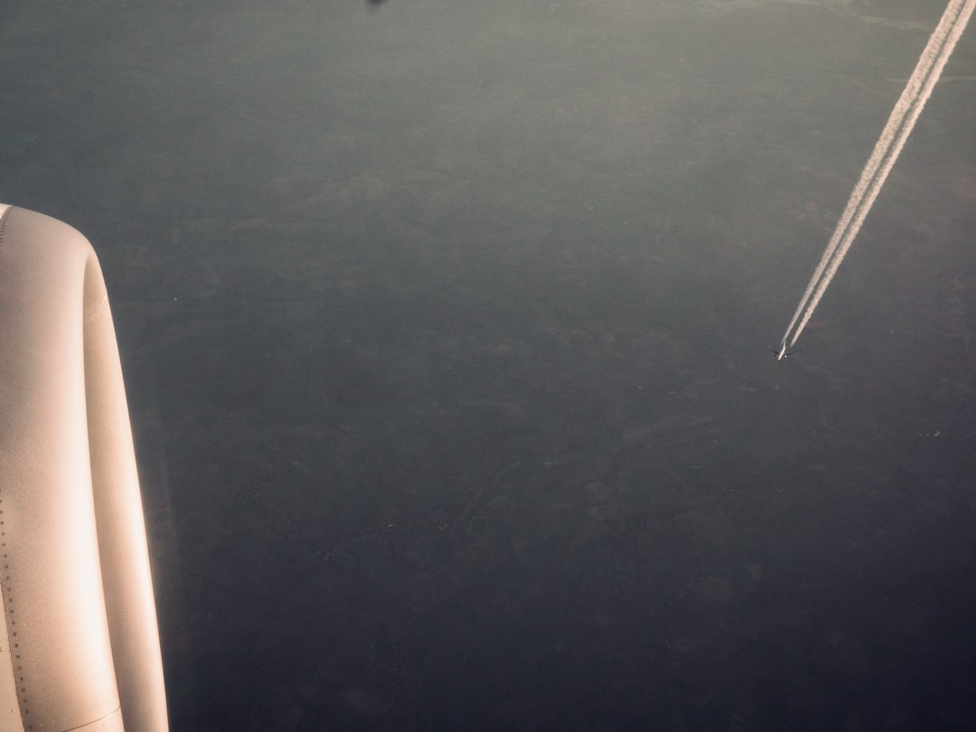 View from an airplane window showing part of the engine on the left and another distant plane on the right, leaving a white contrail against the dark sky below.