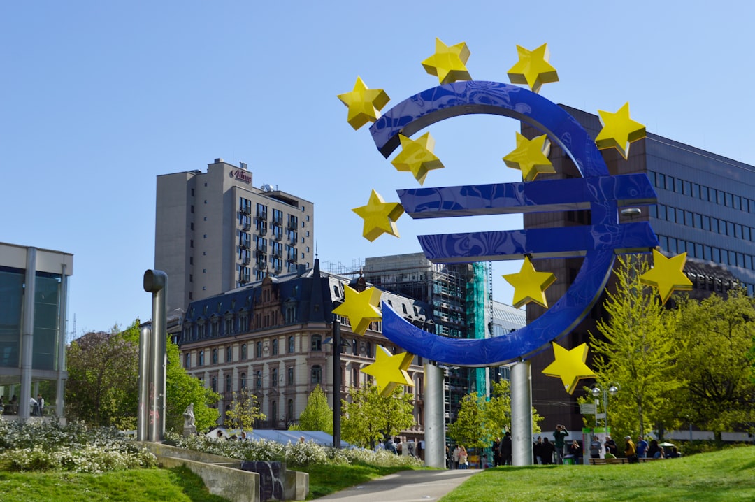 Large blue euro symbol sculpture with yellow stars stands in a city park, surrounded by modern and historic buildings and green trees under a clear blue sky.