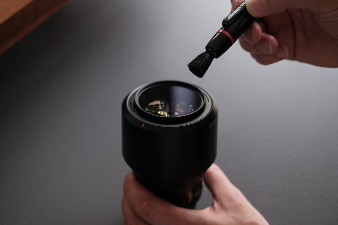 A person uses a small brush to clean the surface of a camera lens, holding the lens steady with one hand on a dark, smooth surface.