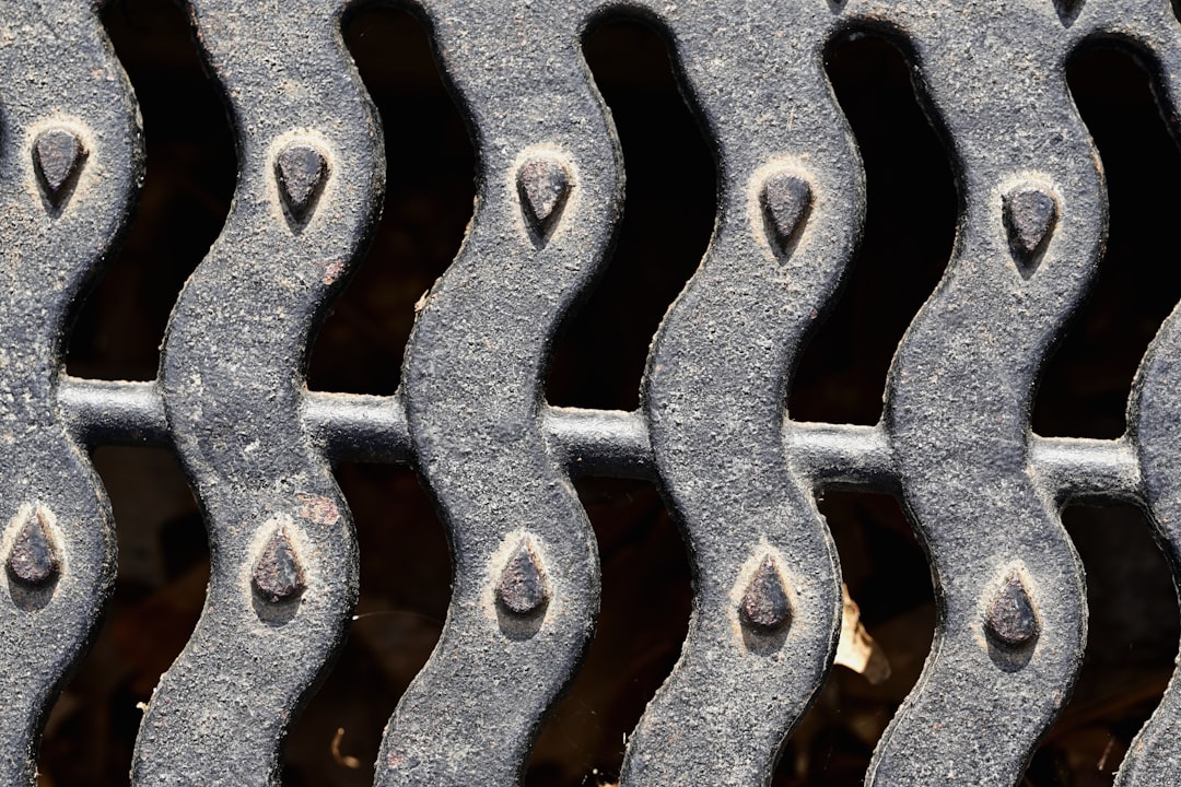 Close-up of a metal grate with a wavy, repeating pattern and teardrop-shaped details on each curve. The surface appears weathered and slightly dusty, with dark gaps between the raised metal sections.