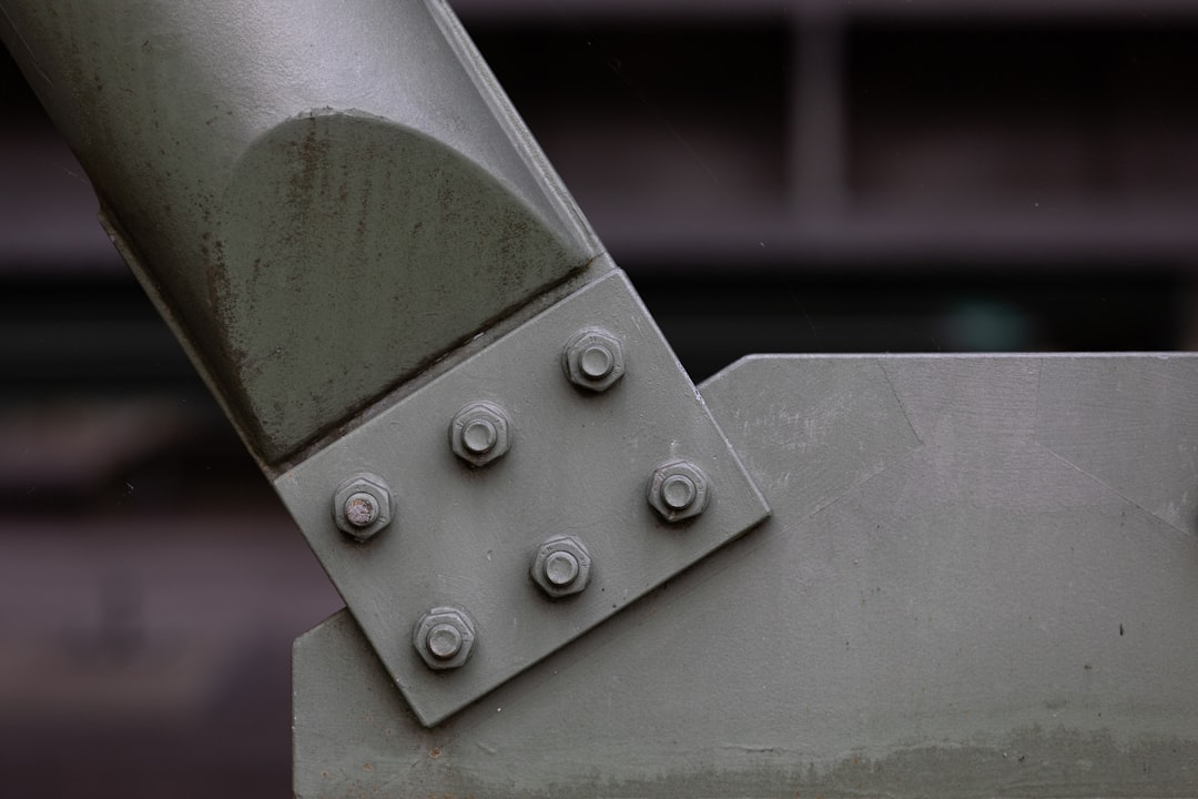 Close-up of a metal structure showing six bolts fastening two gray beams together, with industrial background out of focus.
