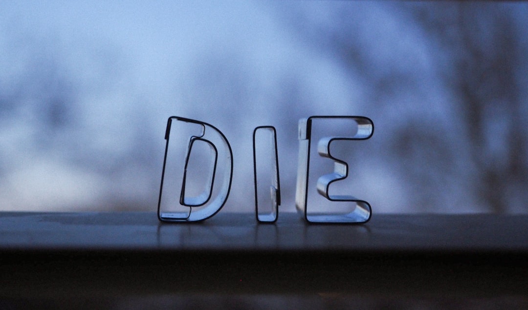 Glass letters spelling "DIE" are placed on a window ledge, with an out-of-focus blue and gray background of trees and sky outside the window.