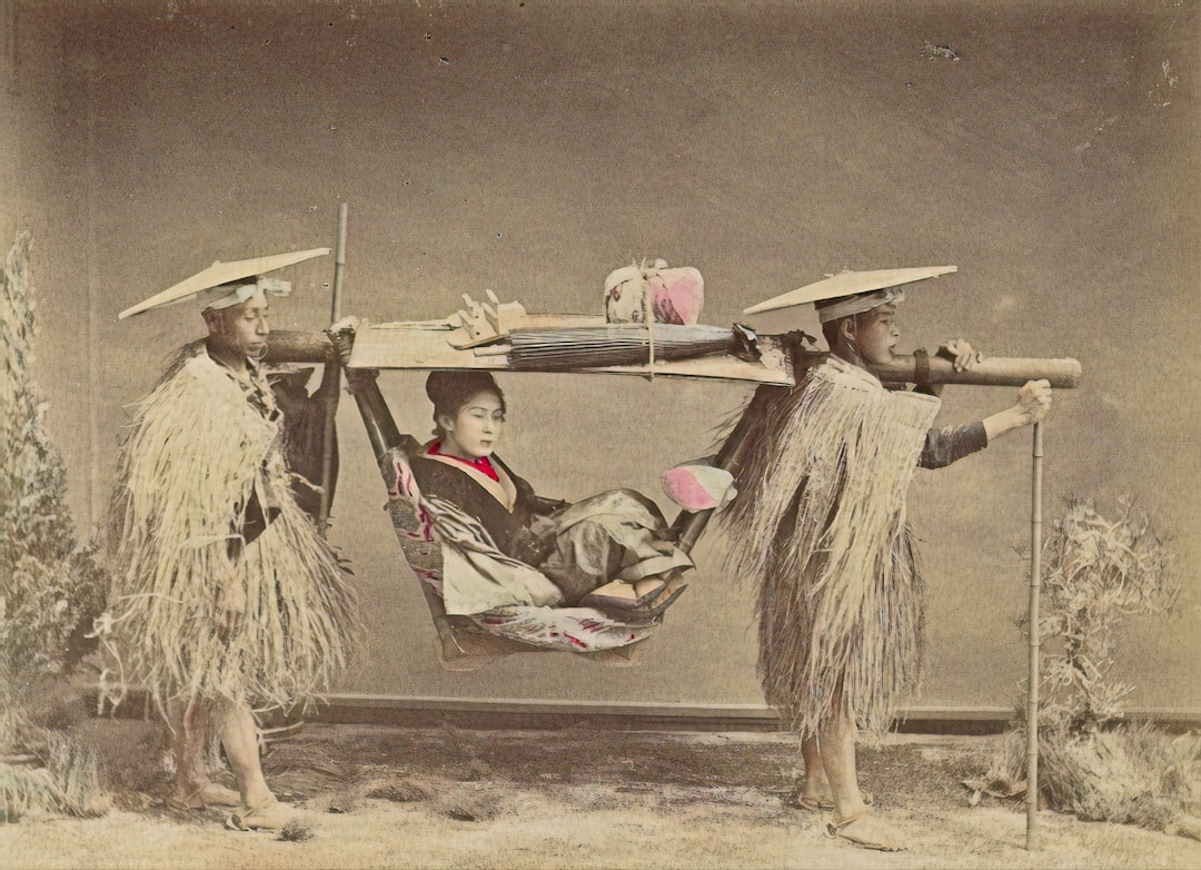 A vintage photo of two men in traditional straw clothing carrying a woman in a palanquin. The woman, dressed in elaborate attire, sits inside while the men use wooden poles to lift and transport the palanquin.