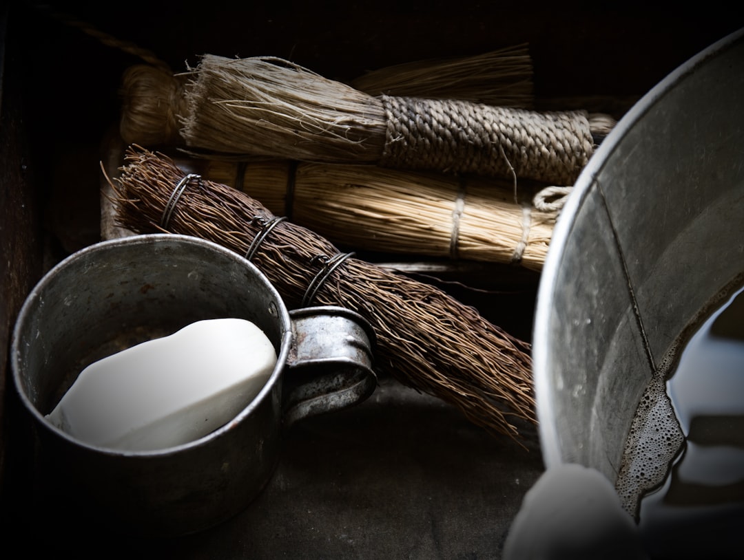 A tin cup holding a bar of white soap sits beside a metal bucket filled with water and several rustic, natural fiber scrub brushes.