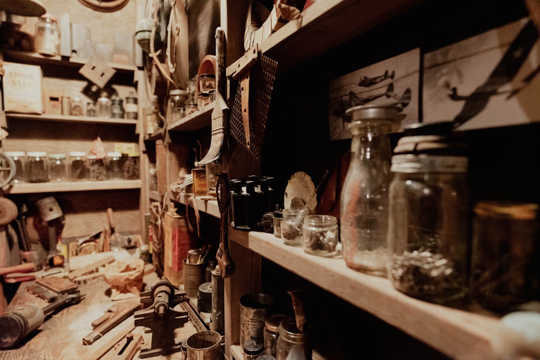 A cluttered workshop with shelves full of jars, tools, and various vintage items, including photos of old airplanes. The scene appears rustic and dimly lit, with workbenches and materials scattered around.