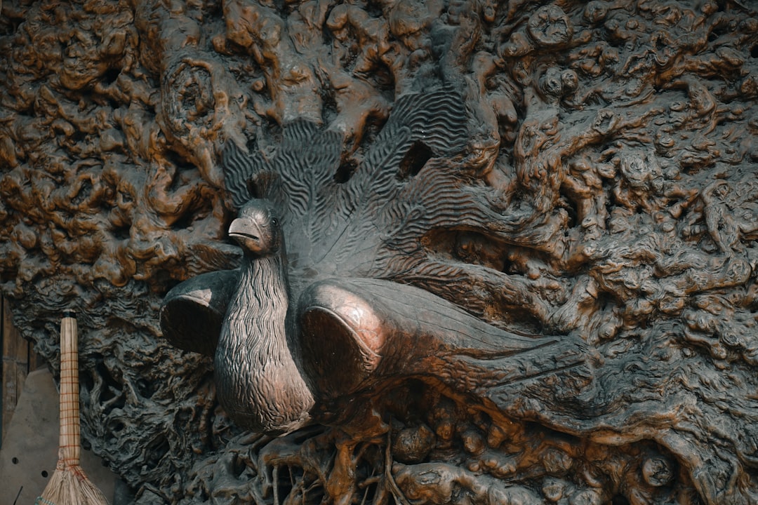 A bronze peacock sculpture is mounted on an intricately textured wall. The detailed background features swirling patterns and adds depth to the artwork. A broom is visible to the left of the sculpture.
