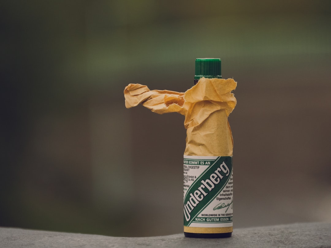 A small green bottle of Underberg herbal digestif stands upright, partially wrapped in crumpled brown paper, against a blurred outdoor background.