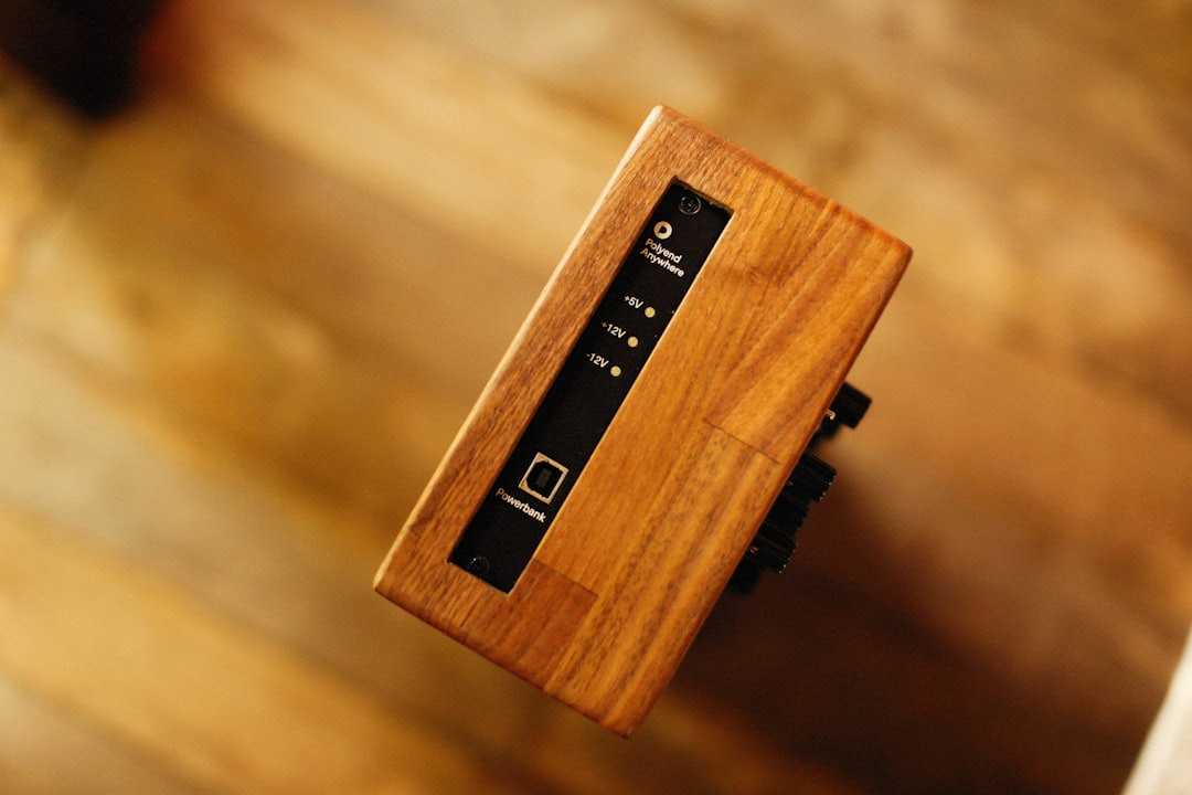 A close-up of a wooden electronic device with a small black control panel and indicator lights, positioned against a blurred wooden background.