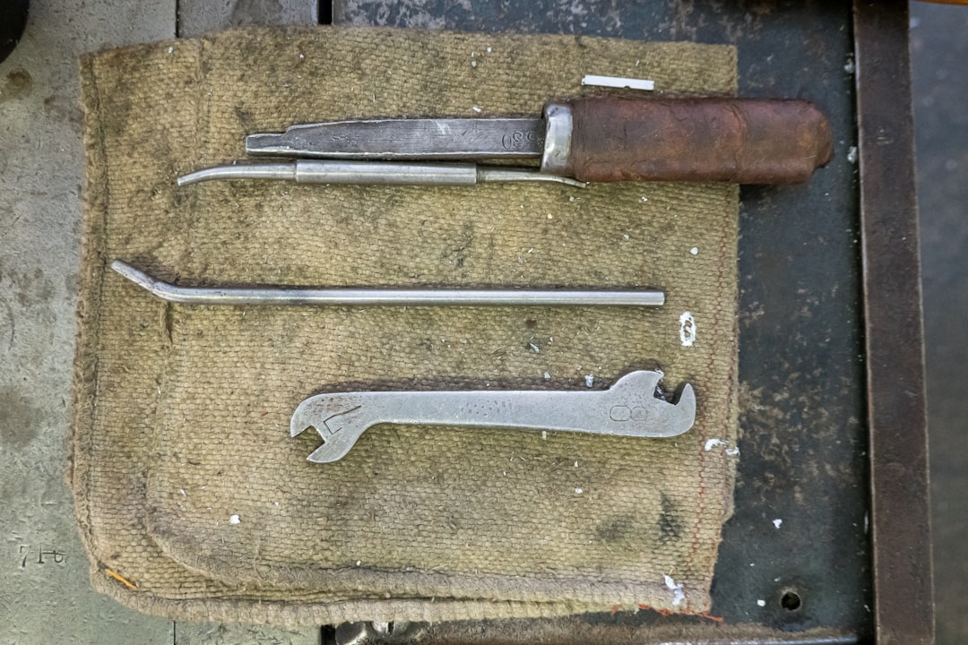 Three vintage metal tools, including a screwdriver with a worn brown handle, a bent rod, and a double-ended wrench, are laid out on a textured, tan cloth on a workbench.
