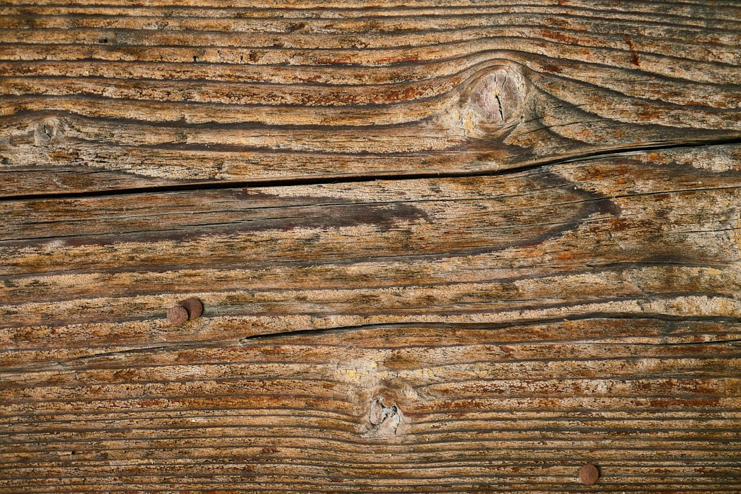 Close-up of weathered wooden boards with visible knots, grain patterns, cracks, and a few rusty nails embedded in the surface. The wood appears old, rough, and textured.