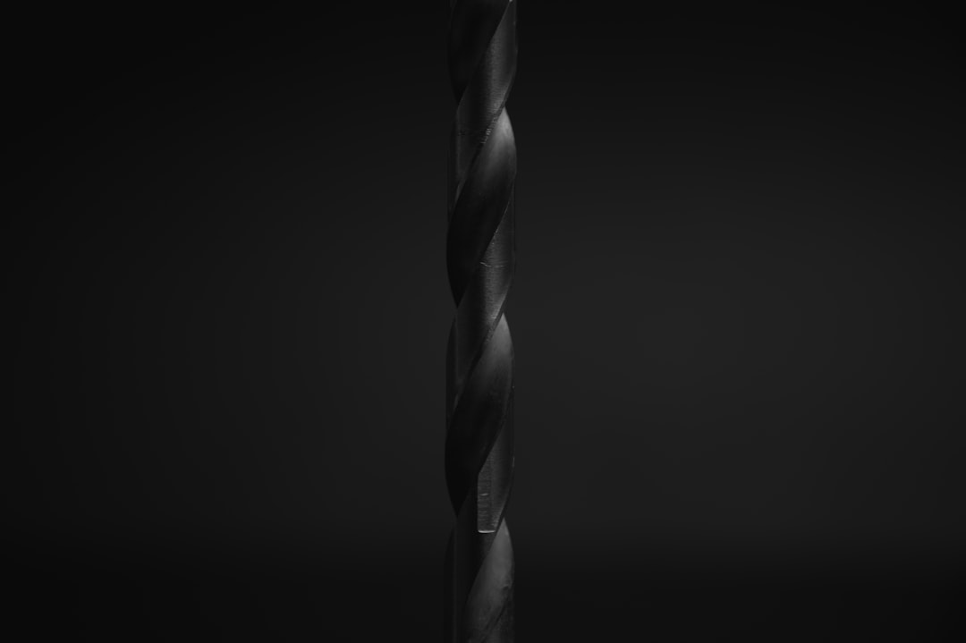 A close-up of a dark, metallic drill bit is shown vertically against a black background, highlighting its spiral grooves and smooth texture with dramatic lighting.