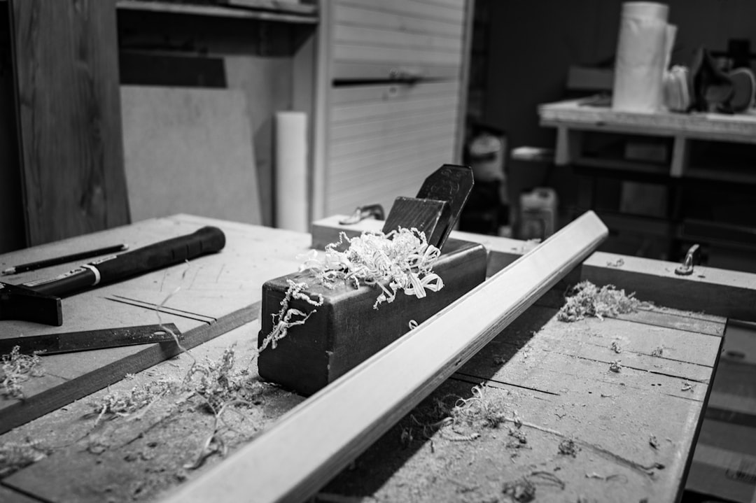 A hand planer with wood shavings rests on a workbench in a carpentry workshop. Tools and materials are visible in the background. The image is in black and white.