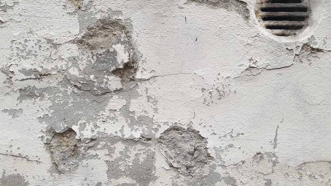 A close-up of an old, weathered wall with peeling, cracked white paint and rough, exposed patches. A metal vent with horizontal slats appears in the top right corner.