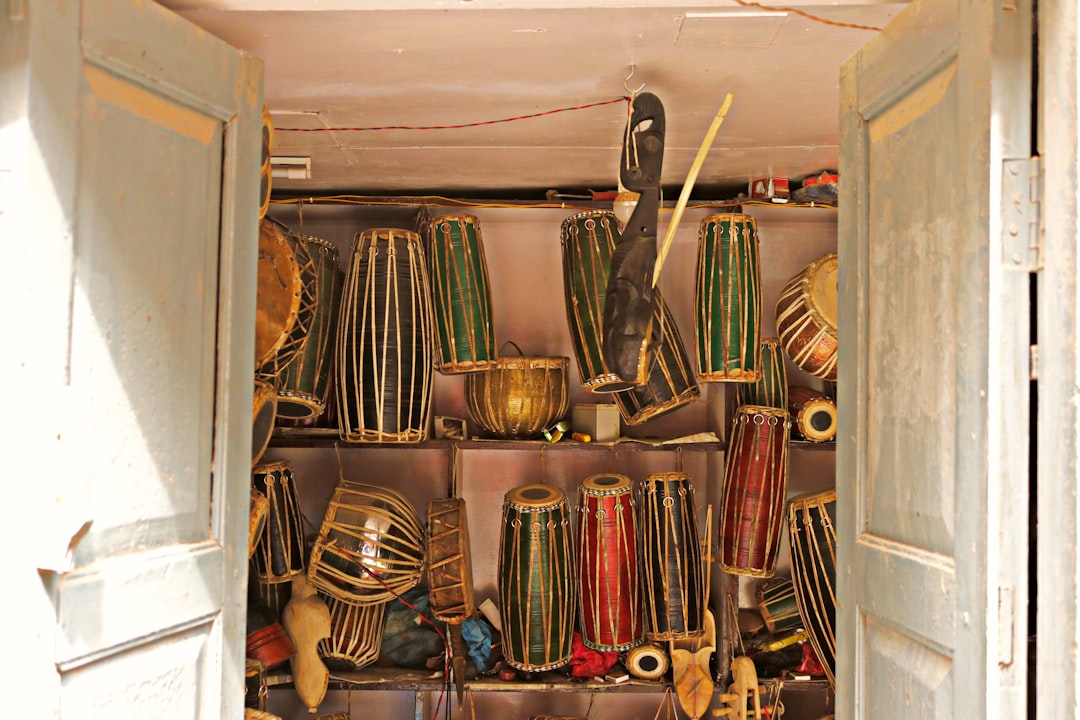 A collection of traditional Indian drums and percussion instruments stacked on shelves inside a small room with worn blue doors slightly open.