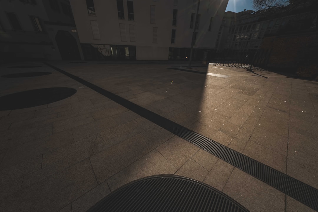 A mostly shadowed urban plaza with tile pavement, intersected by a strip of bright sunlight. The sunlight forms a diagonal pattern across the ground, highlighting the geometric shapes.