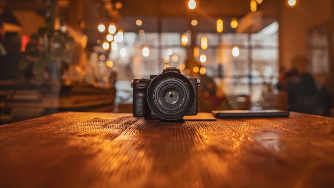A camera sits on a wooden table in a warmly lit café, with blurred lights and people in the background, creating a cozy atmosphere.