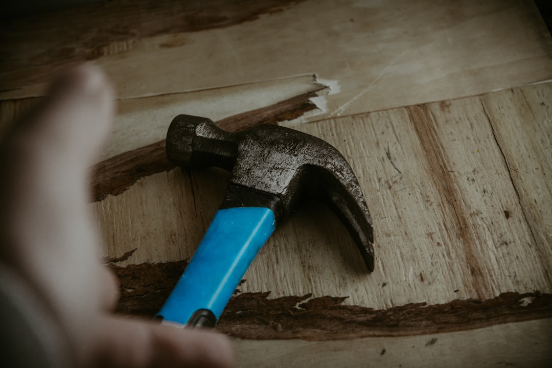 A close-up of a hammer with a blue handle lying on damaged wooden boards, with a blurred hand reaching toward it from the left side of the image.