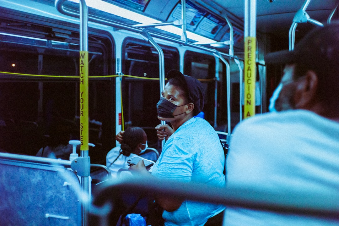 A person wearing a face mask sits on a bus at night, looking toward the camera. Other passengers are seated nearby. The bus interior is lit with blue lighting, and yellow poles display safety warnings.