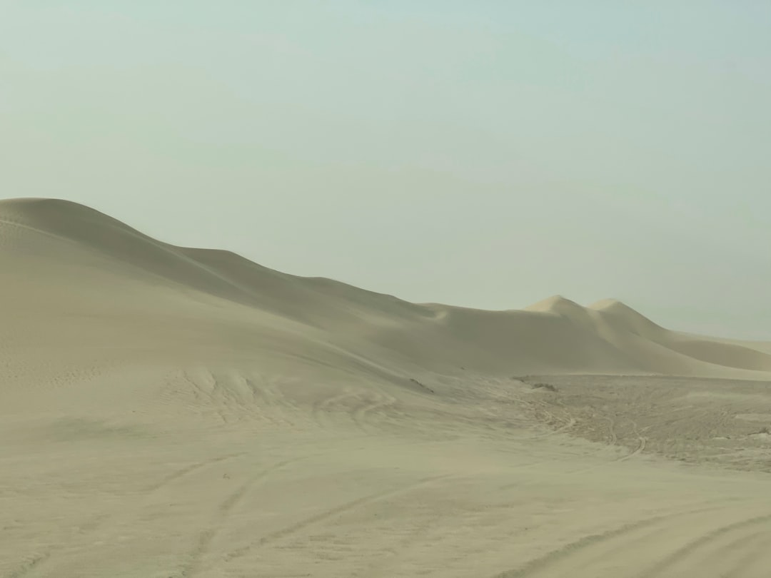 A vast, sandy desert landscape with smooth, rolling sand dunes under a hazy sky. Tire tracks are faintly visible across the foreground.