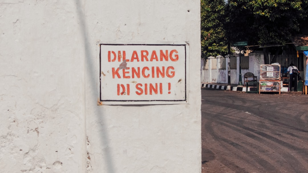 A white wall with a sign in Indonesian that reads "Dilarang Kencing di Sini!" in red letters, meaning "No urinating here!" A street and some people are visible in the background.