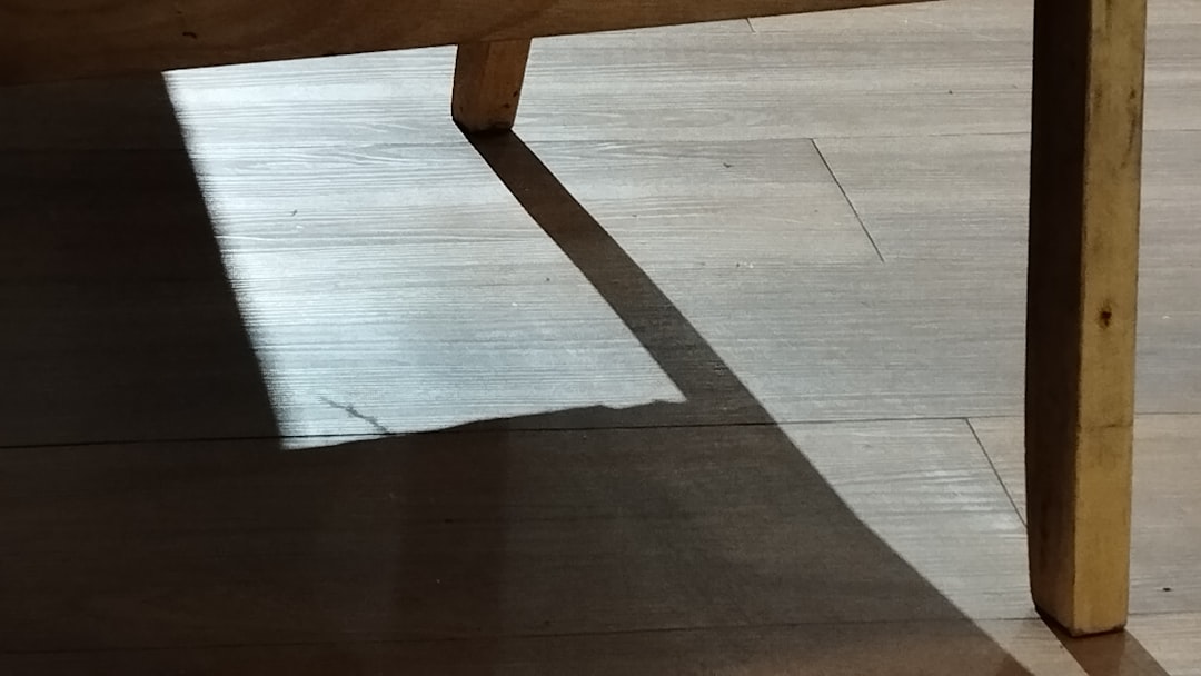 Sunlight casts a sharp-edged shadow of a wooden table leg onto a wooden floor, creating geometric shapes and highlighting the texture of the wood.