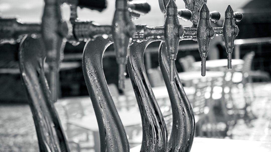 Close-up, black-and-white photo of several shiny metal beer taps in a row, with water droplets on them. Blurry tables and chairs are visible in the background.