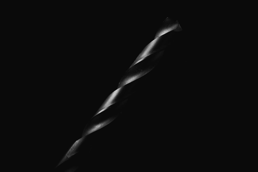 A close-up photo of a metallic drill bit, highlighted by dramatic lighting against a dark, black background, showcasing its twisted, spiral shape.