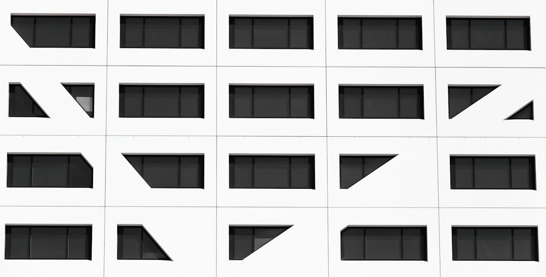 A white building facade with a grid pattern of rectangular black windows, some surrounded by geometric shapes like triangles and slants, creating a modern, abstract appearance.