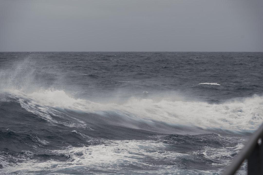 Gray, overcast sky above a choppy, dark ocean with white-capped waves and mist. The water appears rough, and a metal railing is visible in the lower right corner of the image.