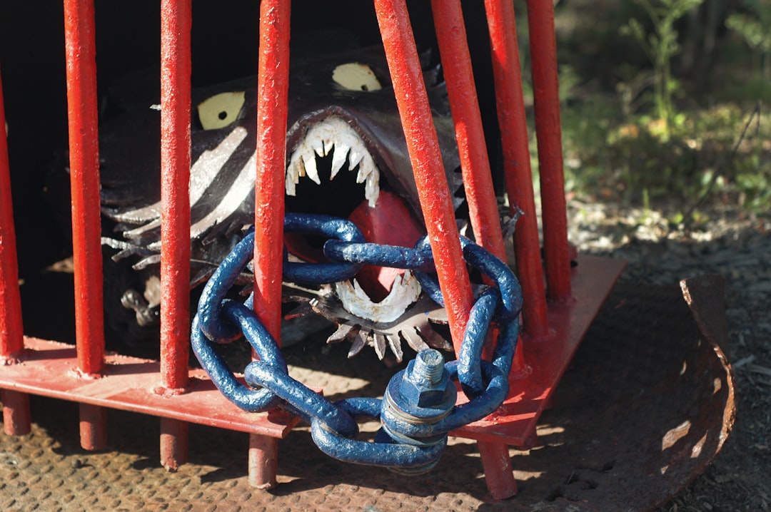 A fierce, black monster with sharp teeth and yellow eyes is behind red metal bars, biting a thick blue chain that secures the cage shut. Sunlight highlights its menacing expression.