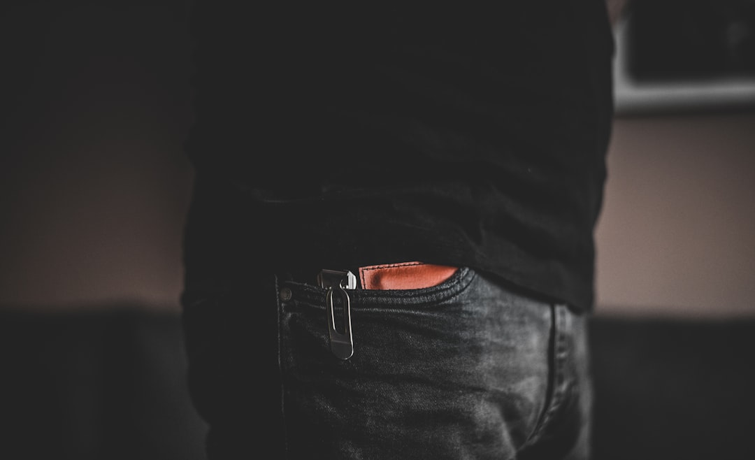 Close-up of a person wearing dark jeans and a black shirt, with a pocket knife clipped to the front right pocket. The background is blurred and out of focus.