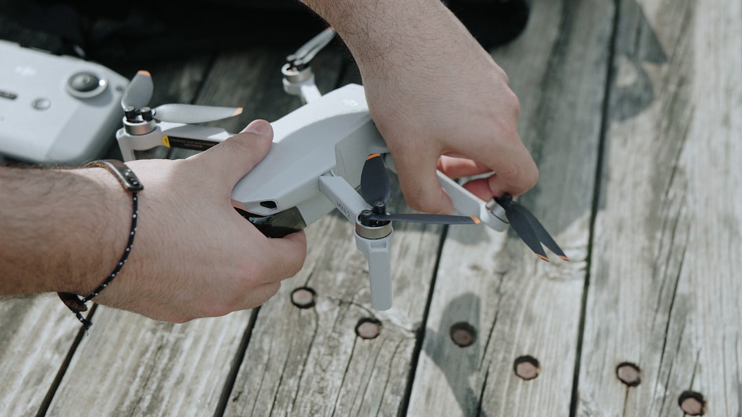 A person is holding a small white drone with both hands on a weathered wooden surface, adjusting its propellers. A drone controller is partially visible beside them.