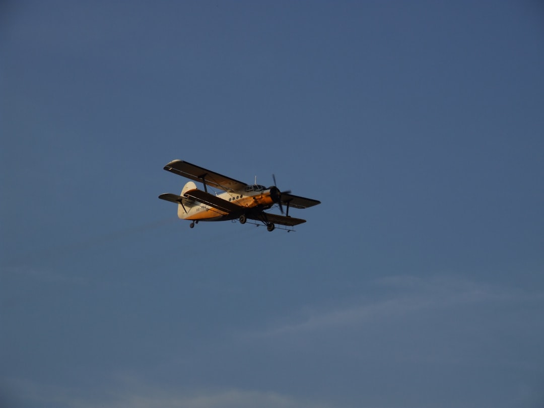 A yellow and white biplane with a propeller flies against a clear blue sky, trailing a faint line of exhaust.
