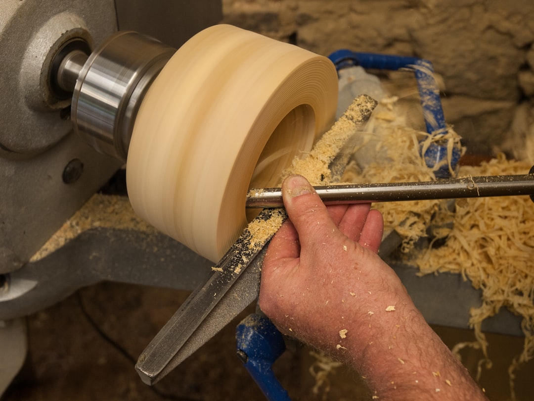 A person uses a chisel to carve a spinning block of wood on a lathe, with wood shavings scattered around.
